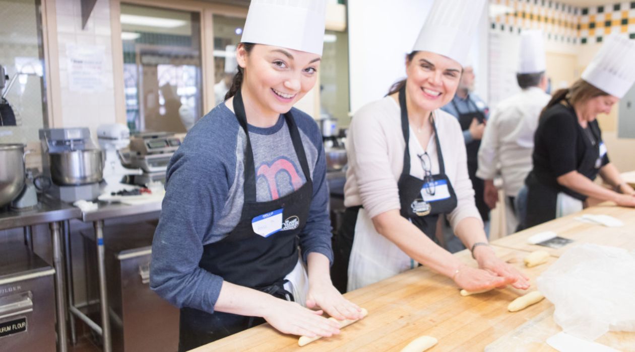 Two female culinary students, wearing tall white chef hats, roll dough on a wooden countertop at the Culinary Institute of America in Hyde Park